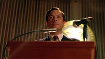 Movie still from “Narcos” (2015), created by Chris Brancato – A man standing at a podium in front of microphones; Medium shot, Low angle