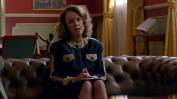 Movie still from “Narcos” (2015), created by Chris Brancato – An older woman sitting on top of a brown couch; Medium shot, Over the shoulder angle