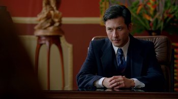 Movie still from “Narcos” (2015), created by Chris Brancato – A man sitting at a desk wearing a suit and tie; Close Up shot, Over the shoulder angle