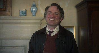Movie still from “Men” (2022), directed by Alex Garland – A man smiling for the camera while wearing a jacket and tie; Medium shot, Over the shoulder angle