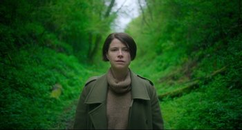 Movie still from “Men” (2022), directed by Alex Garland – A woman wearing a green jacket and a brown scarf; Medium shot, Low angle