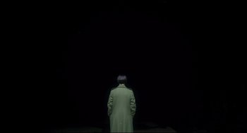 Movie still from “Men” (2022), directed by Alex Garland – A man in a green coat standing in front of a black background; Wide shot, High angle