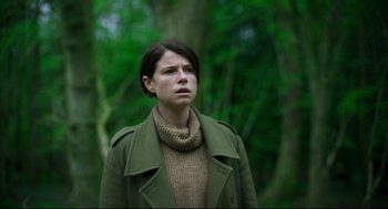 Movie still from “Men” (2022), directed by Alex Garland – A woman wearing a green jacket and a brown sweater; Medium shot, Low angle