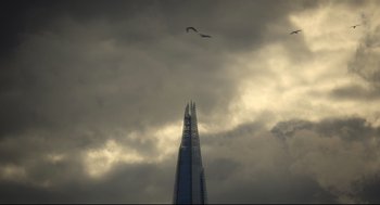 Movie still from “Men” (2022), directed by Alex Garland – A bird flying over a tall building under a cloudy sky; Extreme Wide shot, Low angle