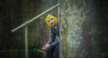 Movie still from “Men” (2022), directed by Alex Garland – A woman with yellow hair sitting on a railing next to a tree; Medium shot, Low angle