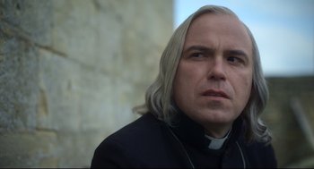 Movie still from “Men” (2022), directed by Alex Garland – A man with long white hair wearing a priest's outfit; Close Up shot, Low angle