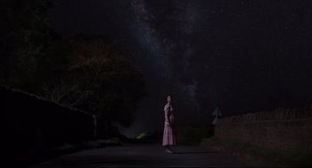 Movie still from “Men” (2022), directed by Alex Garland – A woman standing on the side of a road at night; Extreme Wide shot, Low angle