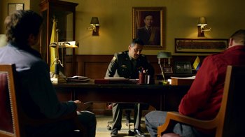 Movie still from “Narcos” (2015), created by Chris Brancato – A man sitting at a table in front of a painting; Medium shot, Over the shoulder angle