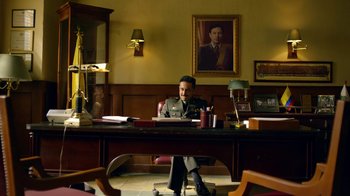 Movie still from “Narcos” (2015), created by Chris Brancato – A man sitting at a desk in front of a painting; Wide shot, Low angle
