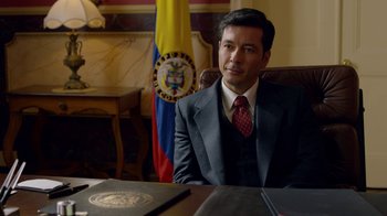 Movie still from “Narcos” (2015), created by Chris Brancato – A man sitting at a desk in front of an american flag; Medium shot, Over the shoulder angle