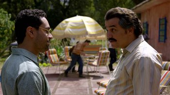 Movie still from “Narcos” (2015), created by Chris Brancato – Two men standing next to each other on a patio; Medium shot, Over the shoulder angle