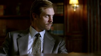 Movie still from “Narcos” (2015), created by Chris Brancato – A man in a suit and tie sitting in front of a bookshelf; Close Up shot, Low angle