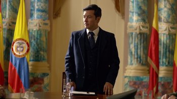 Movie still from “Narcos” (2015), created by Chris Brancato – A man standing at a table with a glass of water; Medium shot, Low angle