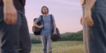 Movie still from “Midsommar” (2019), directed by Ari Aster – A man walking through a field with two bags; Medium shot, Over the shoulder angle