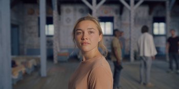 Movie still from “Midsommar” (2019), directed by Ari Aster – A woman in a tan shirt is standing in front of a building; Close Up shot, Over the shoulder angle