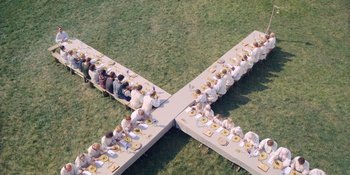 Movie still from “Midsommar” (2019), directed by Ari Aster – A group of people sitting at a long table in the middle of a field; Extreme Wide shot, Overhead angle