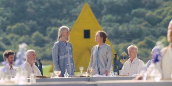 Movie still from “Midsommar” (2019), directed by Ari Aster – Two men standing next to each other in front of a yellow pyramid; Wide shot, Over the shoulder angle