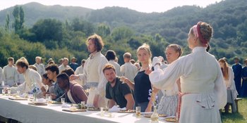 Movie still from “Midsommar” (2019), directed by Ari Aster – A group of people standing around a long table; Wide shot, Over the shoulder angle