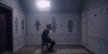 Movie still from “Midsommar” (2019), directed by Ari Aster – A man sitting on a chair in front of a wall; Wide shot, Low angle