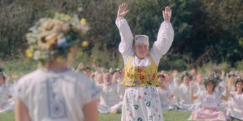 Movie still from “Midsommar” (2019), directed by Ari Aster – An older woman in a floral dress is raising her hands in front of a group of people; Medium shot, Low angle