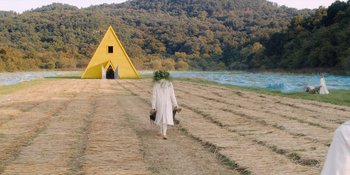 Movie still from “Midsommar” (2019), directed by Ari Aster – A person walking on a dirt road near a yellow pyramid; Extreme Wide shot, Low angle