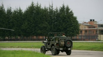 Movie still from “Narcos” (2015), created by Chris Brancato – A man riding in the back of an army jeep; Wide shot, Over the shoulder angle