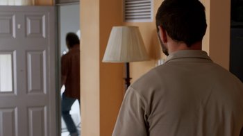 Movie still from “Narcos” (2015), created by Chris Brancato – A man standing next to a lamp in a living room; Medium shot, Over the shoulder angle