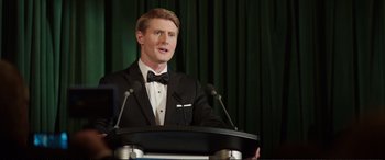 Movie still from “Miss Sloane” (2016), directed by John Madden – A man in a tuxedo standing at a podium with microphones; Medium shot, Low angle