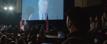 Movie still from “Miss Sloane” (2016), directed by John Madden – A man in front of a large screen with an american flag in front of him; Wide shot, Over the shoulder angle