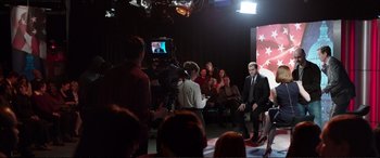 Movie still from “Miss Sloane” (2016), directed by John Madden – A man sitting in front of an audience on a television set; Wide shot, High angle