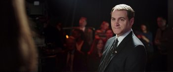 Movie still from “Miss Sloane” (2016), directed by John Madden – A man wearing a suit and tie in front of a group of people; Medium shot, High angle