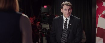 Movie still from “Miss Sloane” (2016), directed by John Madden – A man wearing a suit and tie standing in front of a camera; Medium shot, Over the shoulder angle