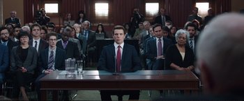 Movie still from “Miss Sloane” (2016), directed by John Madden – A man sitting in front of a group of people in a courtroom; Wide shot, Over the shoulder angle