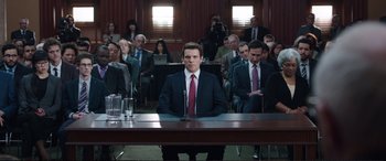 Movie still from “Miss Sloane” (2016), directed by John Madden – A man in a suit and tie sitting in front of a group of people; Wide shot, High angle
