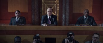 Movie still from “Miss Sloane” (2016), directed by John Madden – A man sitting at a desk in front of two other men; Medium shot, Low angle
