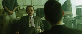 Movie still from “Night in Paradise” (2020), directed by Park Hoon-jung – A man in a suit and tie sitting in front of another man in a suit and tie; Medium shot, Low angle