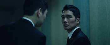 Movie still from “Night in Paradise” (2020), directed by Park Hoon-jung – A man in a suit and tie standing next to another man in a suit and tie; Close Up shot, Over the shoulder angle