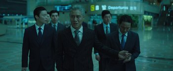 Movie still from “Night in Paradise” (2020), directed by Park Hoon-jung – A group of men in suits and ties walking through an airport terminal; Medium shot, Low angle