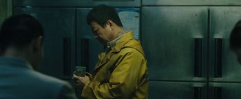 Movie still from “Night in Paradise” (2020), directed by Park Hoon-jung – A man in a yellow jacket looking at his cell phone; Medium shot, Low angle