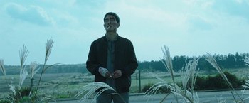 Movie still from “Night in Paradise” (2020), directed by Park Hoon-jung – A man smoking a cigarette while standing next to tall grass; Medium shot, Low angle