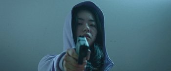 Movie still from “Night in Paradise” (2020), directed by Park Hoon-jung – A woman holding a gun while wearing a hooded sweatshirt; Close Up shot, Low angle