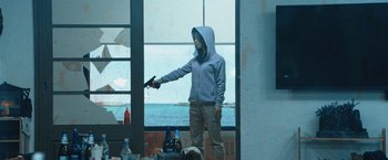 Movie still from “Night in Paradise” (2020), directed by Park Hoon-jung – A person holding a gun while standing in front of a window; Medium shot, High angle