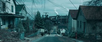 Movie still from “No One Gets Out Alive” (2021), directed by Santiago Menghini – A street with a factory in the background and trees in the foreground; Extreme Wide shot, High angle