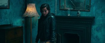Movie still from “No One Gets Out Alive” (2021), directed by Santiago Menghini – A woman in black jacket standing next to a fireplace; Medium shot, Low angle