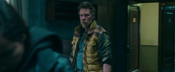 Movie still from “No One Gets Out Alive” (2021), directed by Santiago Menghini – A man in a yellow vest is standing in a dark room; Medium shot, Over the shoulder angle