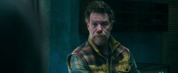 Movie still from “No One Gets Out Alive” (2021), directed by Santiago Menghini – A man with a goatee wearing a plaid jacket; Close Up shot, Over the shoulder angle