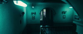 Movie still from “No One Gets Out Alive” (2021), directed by Santiago Menghini – A person standing on a staircase in a room; Wide shot, Low angle