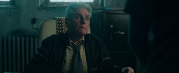 Movie still from “No One Gets Out Alive” (2021), directed by Santiago Menghini – An older man wearing a sweater and a tie; Close Up shot, Low angle