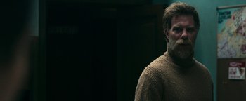 Movie still from “No One Gets Out Alive” (2021), directed by Santiago Menghini – A man with a beard wearing a brown sweater; Close Up shot, Over the shoulder angle