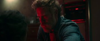 Movie still from “No One Gets Out Alive” (2021), directed by Santiago Menghini – A man with a beard is sitting in a dark room; Close Up shot, Over the shoulder angle
